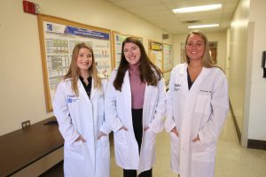 Three female cytotechnology standing in a hallway wearing white lab coats.