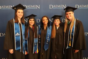 Five female graduates in cap and gowns standing infront of a Daemen backdrop smiling