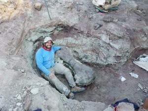 Dominic D’Amore sitting in a hole at a dig site with a dinosaur fossile