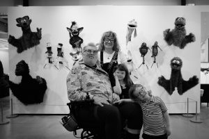 Adam Kreutinger and family in gallery with puppets mounted on wall behind them