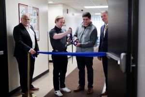 Four men standing in front of a blue ribbon on stanchions with two men in middle holding oversized scissors ready to cut ribbon.