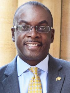 African American man wearing glasses, a white dress shirt, yellow tie and gray suit coat.