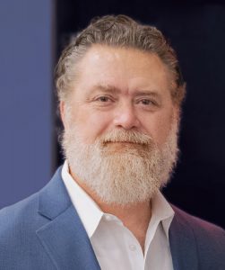 Photo of Dr. Ross wearing a white dress shirt and blue suit coat in front of a black and blue background.