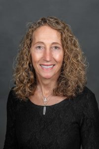 Headshot of a smiling female with long curly light brown hair wearing a black long sleeved shirt and two necklaces