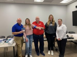 Two males and three females stand in front of a table showing items used in the Stop the Bleed event