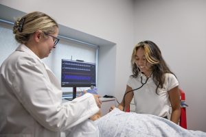 Nursing student with long dark hair and lighter bangs wearing a white polo shirt and using a stethoscope and a faculty member with blonde hair, glasses and wearing a white lab coat practice on a manekin laying in bed in a simulated hospital room.