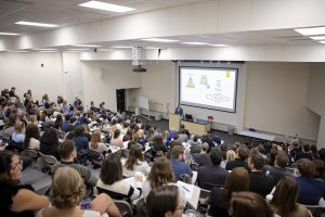 Classroom auditorium filled with PT students with presenter at the front behind a podium and in front of large screen with presentation displayed.