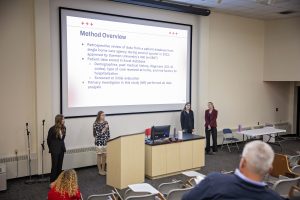 Four female PT students standing in front of lecture auditorium presenting slides on the large screen behind them