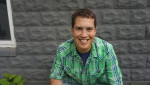 Male dressed in a light green plaid shirt sitting in front of a gray stone house.