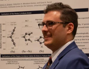 Male professor with short brown hair and glasses wearing a suit and tie standing in front of a chemistry diagram.