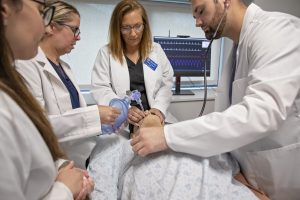 Professor and students dressed in white coats working on a manikin in a simulated hospital room setting.