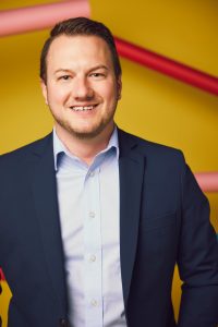 Male with short brown hair dressed in a light blue dress shirt and blue sport coat in front of a yellow wall.