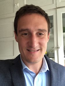 Man with short dark brown hair wearing a light blue collared shirt and dark blue sport coat.