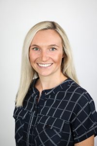 Female with long blonde hair, smiling, wearing a black short sleeved blouse in front of a light gray background.