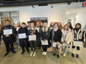 Group of high school students standing in the art gallery in front of a white wall with artwork hanging on it. 