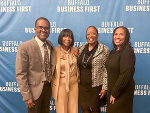 An African American man dressed in a suit with three African American women dressed in professional attire in front of a blue Buffalo Business First backdrop.