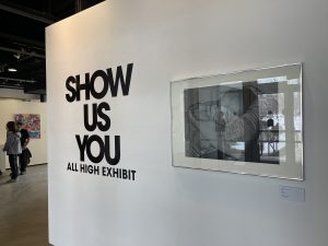 White wall with framed artwork handing on it and "Show us You All High Exhibit" black text on the wall