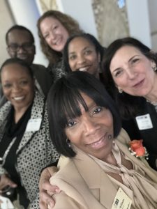 Selfie of an African American woman with short black hair with four other women and a man, all dressed in professional clothing.