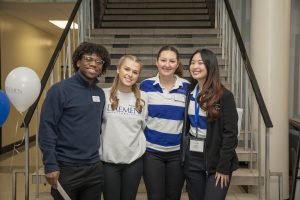 Four students (African American man and three women with long hair standing arm-in-arm standing in front of a staircase. 