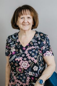 Female with short light brown hair, smiling, wearing a short sleeved floral v-neck blouse standing in front of a light gray background.