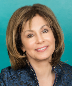 Woman with shoulder-length light brown hair, smiling, wearing a black and blue jacket in front of a teal wall