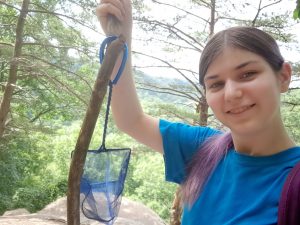 Female with long dark hair in a pony tail holding a small net with an isopod inside with a wooded background.