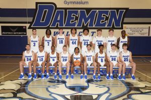 Image of 17 male basketball players in white Daemen uniforms in the gymnasium. Eight men are standing and nine men are sitting.