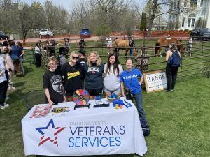 Group of 5 women standing behind a Veterans Services table with horses in a corral on the lawn behind them