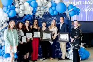 Group of 8 men and women, some of which are holding awards, standing in front of a blue Daemen backdrop and blue and white balloons
