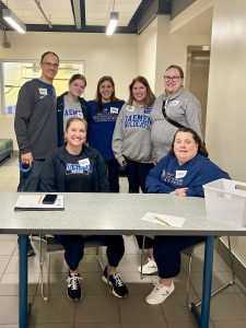 Seven men and women standing and sitting behind a table, wearing Daemen attire. 
