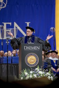 Man wearing glasses and a cap and gown standing behind a Daemen podium with his hands outstretched speaking to an audience.