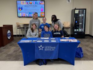 Six women students wearing Daemen shirts standing and sitting behind a display table that says Veteran Services