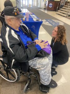 Man in a wheel chair wearing a baseball cap talking to a woman who is crouched down looking at him
