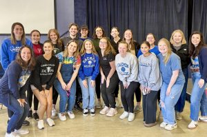 19 men and women standing together smiling in a room, most of which are wearing Daemen sweatshirts