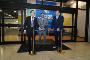 Three men dressed in suits and ties standing behind stanchions and a blue ribbon ready to cut the ribbon
