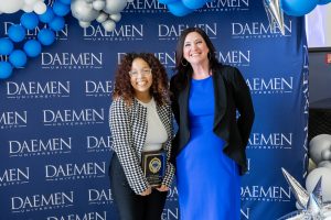 Two women with long brown hair standing in front of a blue Daemen backdrop