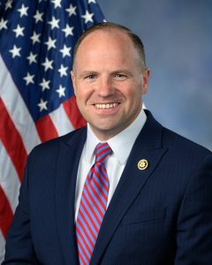 Male wearing a white dress shirt, red and light blue striped tie and navy suit jacket posing in front of an American flag. 