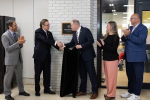 Two men shaking hands in front of a plaque with two man and a woman applauding