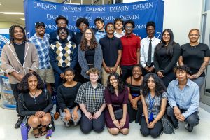 20 male and female students standing and kneeling in front of a blue Daemen backdrop