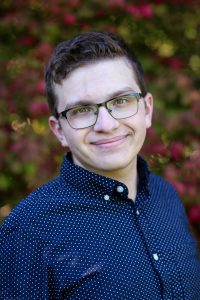 Young man with dark hair and glasses, wearing a navy button down shirt standing in front of flowering plant