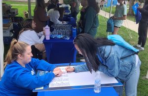 Two women at a table. One with long dark hair wearing a jean jacket standing and writing on a large board on a table. The other with long blond hair pulled in a ponytail wearing a royal blue sweatshirt.