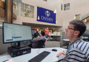 Male sitting at a desk in a library looking at a computer monitor with a blue Daemen flag hanging on the wall in the background
