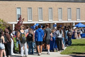 Group of students standing in line on college campus sidewalk with a building in the background