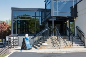 Building with glass windows and stairs leading up to the entrance. Blue sign that says Daemen Hall with blue and white balloons tied to it.