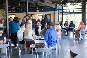 Large room with bistro tables and windows with several people standing and sitting