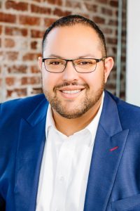Male with short brown hair, a beard and glasses wearing a white dress shirt and a blue blazer, standing in front of a brick wall.