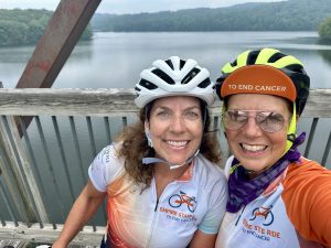Two female cyclists wearing biking jerseys and helmets standing on a bridge with water in the background