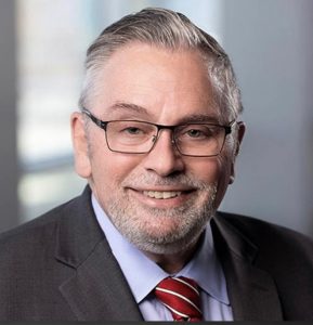 Man with salt and pepper hair, wearing glasses, a light blue dress shirt, striped red tie and dark gray suit jacket
