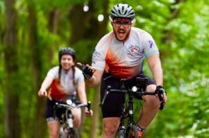 Male cyclist wearing biking shorts, jersey and helmet riding his bike on a trail with trees and a female rider behind him