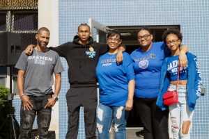 African American family wearing Daemen t-shirts and hoodies standing in front of a residence hall